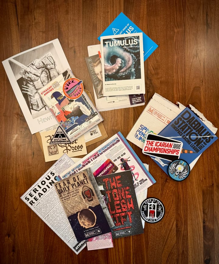 Splay of zines on wood table. They are collected into piles on top of the envelopes they arrived in.